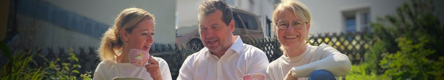 V.l.n.r. Annette Ehrlich, Holger K&ouml;ckritz und Annette Currle sitzen im Garten und genie&szlig;en Tee aus rosa Tassen. Sie tragen wei&szlig;e Oberteile und l&auml;cheln in die Kamera., &copy; Perle Uhlbach