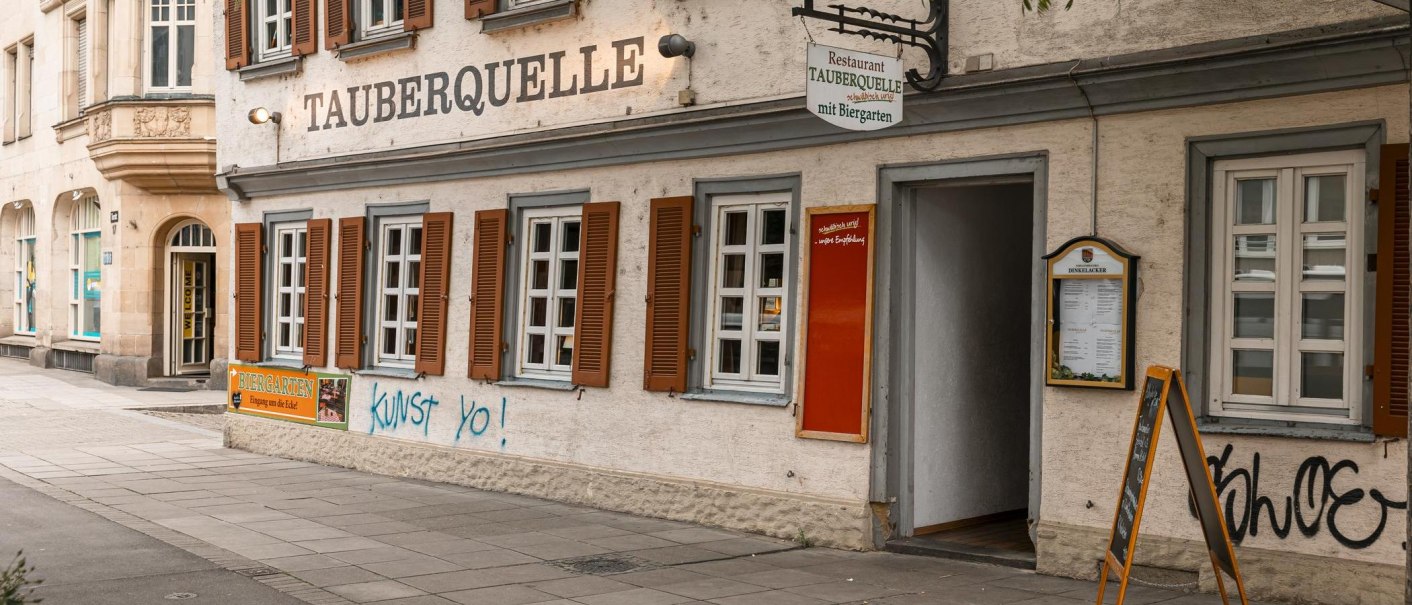 Das Restaurant Tauberquelle in Stuttgart zeigt eine traditionelle Fassade mit Fensterläden und Graffiti. Ein Schild weist auf den Biergarten hin., © SMG, Sarah Schmid