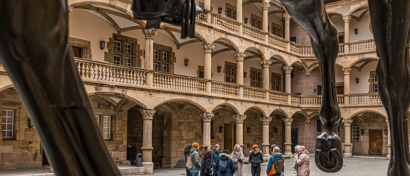 Innenhof eines alten Schlosses mit Arkaden und Säulen. Eine Gruppe von Menschen steht zusammen, im Vordergrund sind die Beine einer Pferdestatue sichtbar., © Stuttgart Marketing GmbH - Sarah Schmid