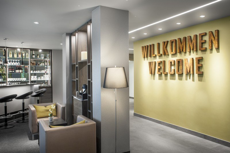 A modern lobby with a bar, a seating area, and a large "Welcome" sign on the wall. Elegant decor featuring bar stools and a floor lamp., &copy; TOMAS