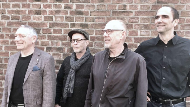 Vier Männer stehen lächelnd vor einer Backsteinmauer und blicken nach rechts. Sie tragen lässige Kleidung und wirken entspannt., © Birgitta Menzer Vier Männer stehen lächelnd vor einer Backsteinmauer und blicken nach rechts. Sie tragen lässige Kleidung und wirken entspannt., © Birgitta Menzer