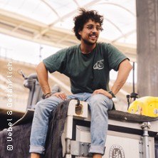 Ein Mann mit lockigem Haar sitzt entspannt auf einem großen Koffer in einer Halle. Er trägt ein grünes T-Shirt und Jeans., © links im Bild Ein Mann mit lockigem Haar sitzt entspannt auf einem großen Koffer in einer Halle. Er trägt ein grünes T-Shirt und Jeans., © links im Bild