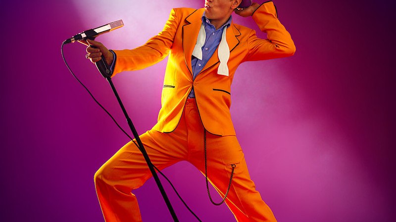 Eine Person in einem auffälligen orangefarbenen Anzug und Hut posiert mit einem Mikrofon vor einem lila Hintergrund. Der Schriftzug "HEROES" ist oben zu sehen., © MSK Events GmbH Eine Person in einem auffälligen orangefarbenen Anzug und Hut posiert mit einem Mikrofon vor einem lila Hintergrund. Der Schriftzug "HEROES" ist oben zu sehen., © MSK Events GmbH