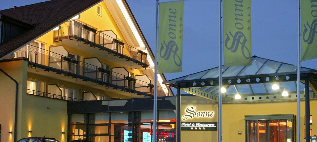 Hotel Sonne at dusk, illuminated entrance, yellow building with balconies, three flags in front of it., © Hotel & Restaurant Sonne