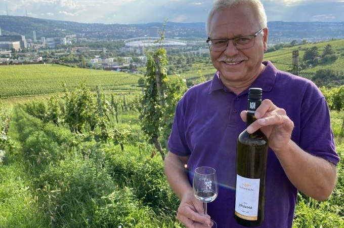 Ein Mann in einem lila Hemd h&auml;lt eine Weinflasche und ein Glas in einem Weinberg. Im Hintergrund ist Stuttgart mit einem Stadion zu sehen., &copy; Weingenuss Stuttgart