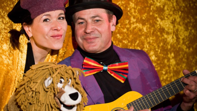 Zwei Personen in bunten Kostümen, eine mit Löwenpuppe, die andere mit Gitarre, vor goldenem Hintergrund. Beide lächeln in die Kamera., © Liedertheater Altmann