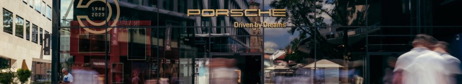 Glass façade of the Porsche Brand Store with anniversary logo '75 years 1948-2023'. People walk past, buildings and trees are reflected in the glass., © Porsche AG