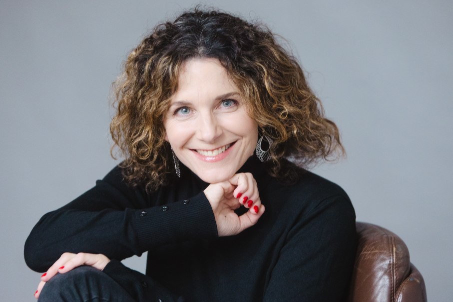 Eine Frau mit lockigem Haar und Ohrringen l&auml;chelt in die Kamera. Sie tr&auml;gt einen schwarzen Pullover und sitzt auf einem braunen Ledersessel., &copy; Uwe Hauth