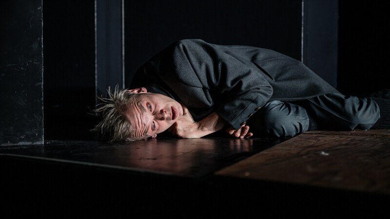 Ein Schauspieler in einem dunklen Raum liegt auf dem Boden und schaut nachdenklich in die Kamera. Die Szene wirkt dramatisch und intensiv., © Württembergische Staatstheater Stuttgart Ein Schauspieler in einem dunklen Raum liegt auf dem Boden und schaut nachdenklich in die Kamera. Die Szene wirkt dramatisch und intensiv., © Württembergische Staatstheater Stuttgart
