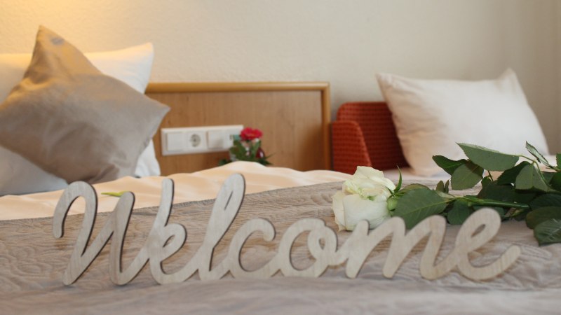Ein Bett mit Kissen, einer weißen Rose und einem 'Welcome'-Schild. Im Hintergrund eine Steckdose und ein roter Sessel., © TOMAS