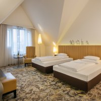 Ein modernes Hotelzimmer mit zwei Einzelbetten, einem Schreibtisch, einem Sessel und einem Flachbildfernseher. Die Einrichtung ist in warmen Tönen gehalten., © ARCOTEL Hotels Ein modernes Hotelzimmer mit zwei Einzelbetten, einem Schreibtisch, einem Sessel und einem Flachbildfernseher. Die Einrichtung ist in warmen Tönen gehalten., © ARCOTEL Hotels