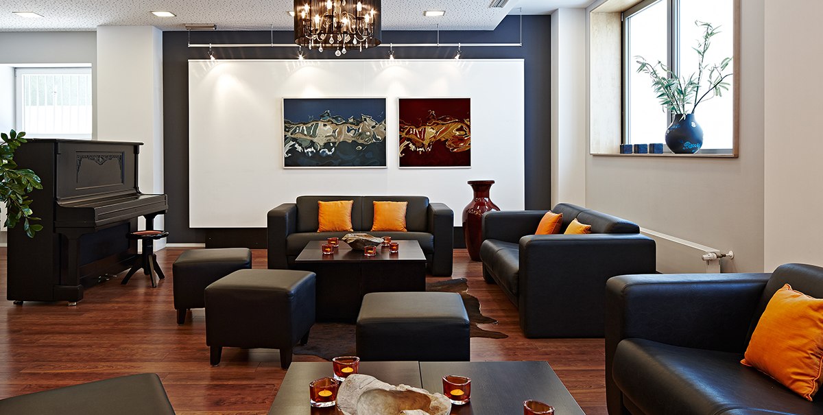 Elegante Lounge mit schwarzen Sofas, orangefarbenen Kissen, Klavier, Kunstwerken und Kerzen auf Holztischen., © Studierendenwerk Stuttgart