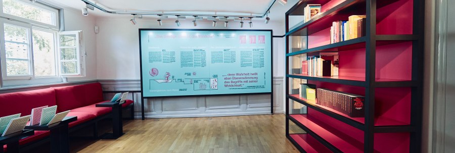 Hegel-Haus Dauerausstellung: Raum mit roten Sitzbänken, einem großen Bücherregal und einer Infotafel an der Wand. Helle, moderne Einrichtung., © Julia Ochs