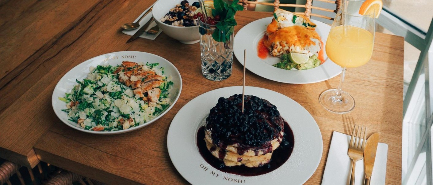 Ein Tisch mit Salat, Pfannkuchen mit Beeren, einem Getränk und weiteren Speisen auf Holztisch., © The Gardeners Nosh, Stuttgart