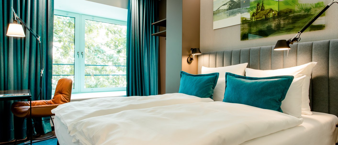 Modern hotel room with large bed, blue pillows and curtains. A window offers a view of the greenery. There is a desk and chair next to it., &copy; Motel One - Stuttgart Hauptbahnhof