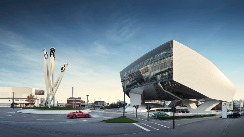 The Porsche Museum in Stuttgart with its futuristic architecture and a sculpture of three cars on tall pillars against a blue sky., &copy; Porsche AG