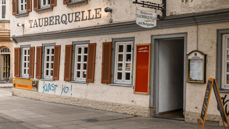 Das Restaurant Tauberquelle in Stuttgart zeigt eine traditionelle Fassade mit Fensterläden und Graffiti. Ein Schild weist auf den Biergarten hin., © SMG, Sarah Schmid Das Restaurant Tauberquelle in Stuttgart zeigt eine traditionelle Fassade mit Fensterläden und Graffiti. Ein Schild weist auf den Biergarten hin., © SMG, Sarah Schmid