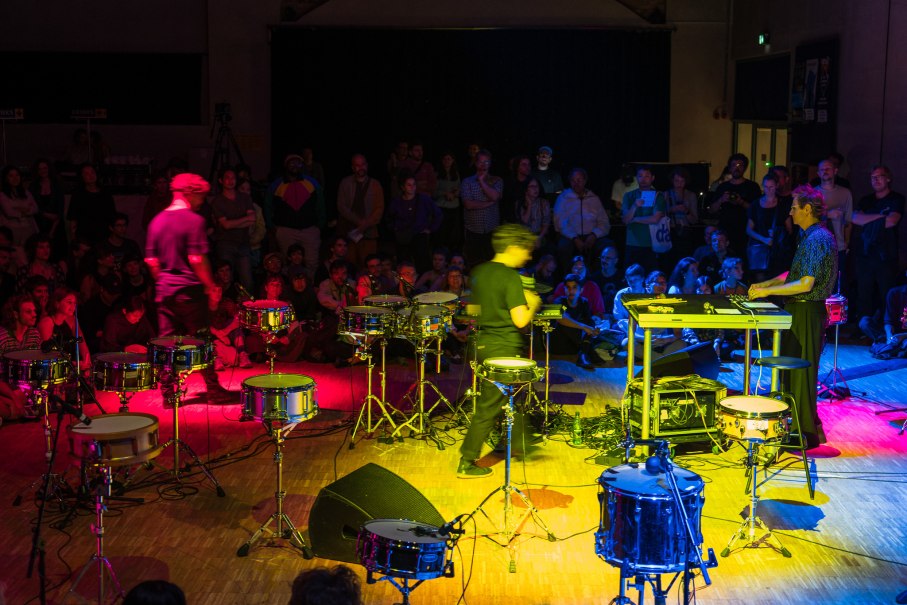 Ein Konzert mit Schlagzeug und elektronischen Instrumenten, umgeben von einer bunten Menschenmenge. Die B&uuml;hne ist in lebhaften Farben beleuchtet., &copy; IMD/Kristof Lemp