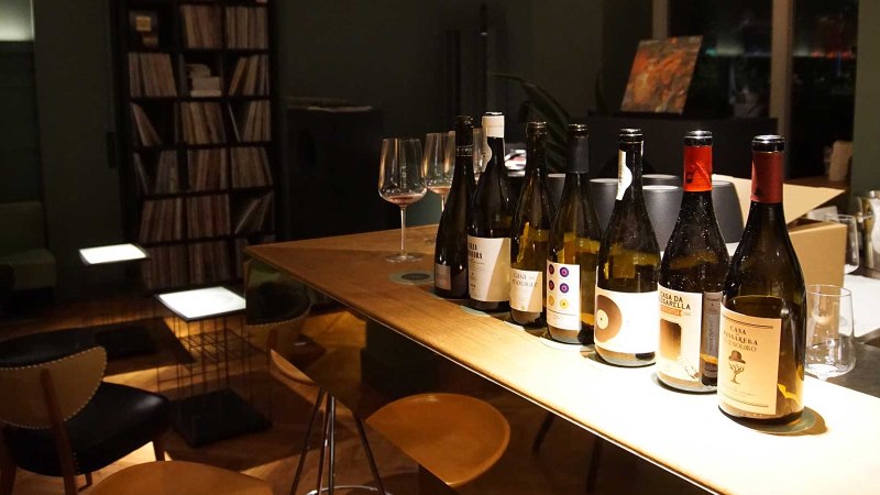 Mehrere Weinflaschen stehen auf einem Holztisch in einer stilvollen Bar. Im Hintergrund sind ein Schallplattenregal und gemütliche Sitzgelegenheiten zu sehen., © Kati Guenter Mehrere Weinflaschen stehen auf einem Holztisch in einer stilvollen Bar. Im Hintergrund sind ein Schallplattenregal und gemütliche Sitzgelegenheiten zu sehen., © Kati Guenter
