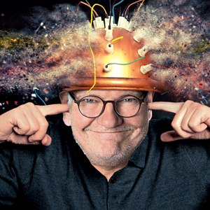 A man wearing glasses is wearing a helmet with wires and smoke coming out of it; he is covering his ears and smiling at the camera., © Renitenztheater Stuttgart e.V. A man wearing glasses is wearing a helmet with wires and smoke coming out of it; he is covering his ears and smiling at the camera., © Renitenztheater Stuttgart e.V.