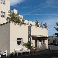 A beige architecture gallery with a flat roof, benches, and a large conifer tree against a blue sky., © Thomas Fütterer A beige architecture gallery with a flat roof, benches, and a large conifer tree against a blue sky., © Thomas Fütterer