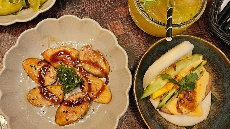 Ein Teller mit veganen Teigtaschen in Soße, ein Bao Bun mit Gemüse und ein Getränk mit Eiswürfeln auf einem Holztisch., © SMG Ein Teller mit veganen Teigtaschen in Soße, ein Bao Bun mit Gemüse und ein Getränk mit Eiswürfeln auf einem Holztisch., © SMG