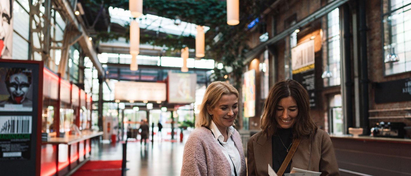 Two women are standing in the foyer of a theater looking at tickets. Posters and modern architecture can be seen in the background., © Stuttgart-Marketing GmbH, wpsteinheisser