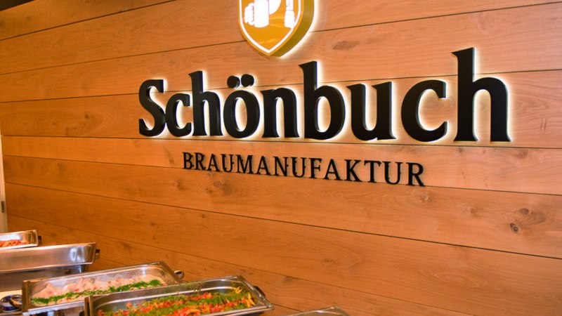 Interior view of a brewery with buffet. "Schönbuch Braumanufaktur" is written on the wooden wall. In the foreground are dishes in warming containers., © © Schönbuchbrauerei, Sebastian Berger
