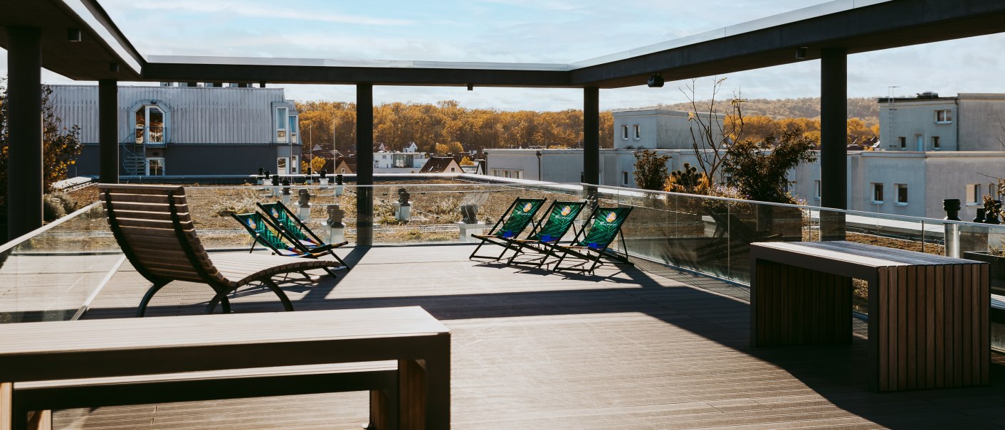 Modern roof terrace with sun loungers and wooden furniture, surrounded by glass railings, with a view of the building and the autumn landscape., © Rioca Stuttgart Posto 4 GmbH Modern roof terrace with sun loungers and wooden furniture, surrounded by glass railings, with a view of the building and the autumn landscape., © Rioca Stuttgart Posto 4 GmbH