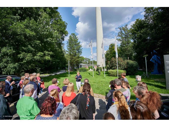 A group of people take part in a guided tour in front of a tall tower outside. The sky is partly cloudy., &copy; SWR Media Services GmbH / Fernsehturm Stuttgart