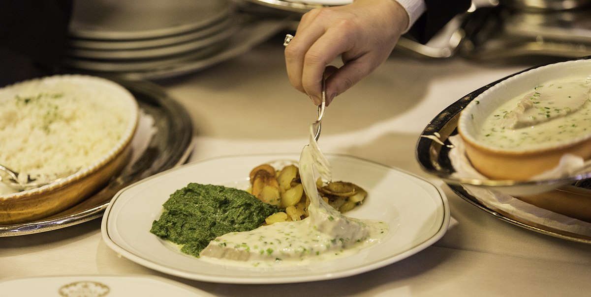 Ein Teller mit Spinat, Bratkartoffeln und einer weißen Sauce wird serviert. Eine Hand hält einen Löffel mit Sauce., © Hotel Royal