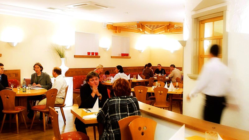 Cozy restaurant with wooden furniture, several guests at tables and a waiter on the move. Warm lighting and decorative elements on the walls., &copy; SMG