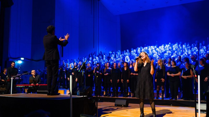 Ein großer Chor singt auf einer Bühne, beleuchtet von blauem Licht. Eine Solistin steht vorne, während ein Dirigent den Chor leitet., © Ludmilla Parsyak Ein großer Chor singt auf einer Bühne, beleuchtet von blauem Licht. Eine Solistin steht vorne, während ein Dirigent den Chor leitet., © Ludmilla Parsyak