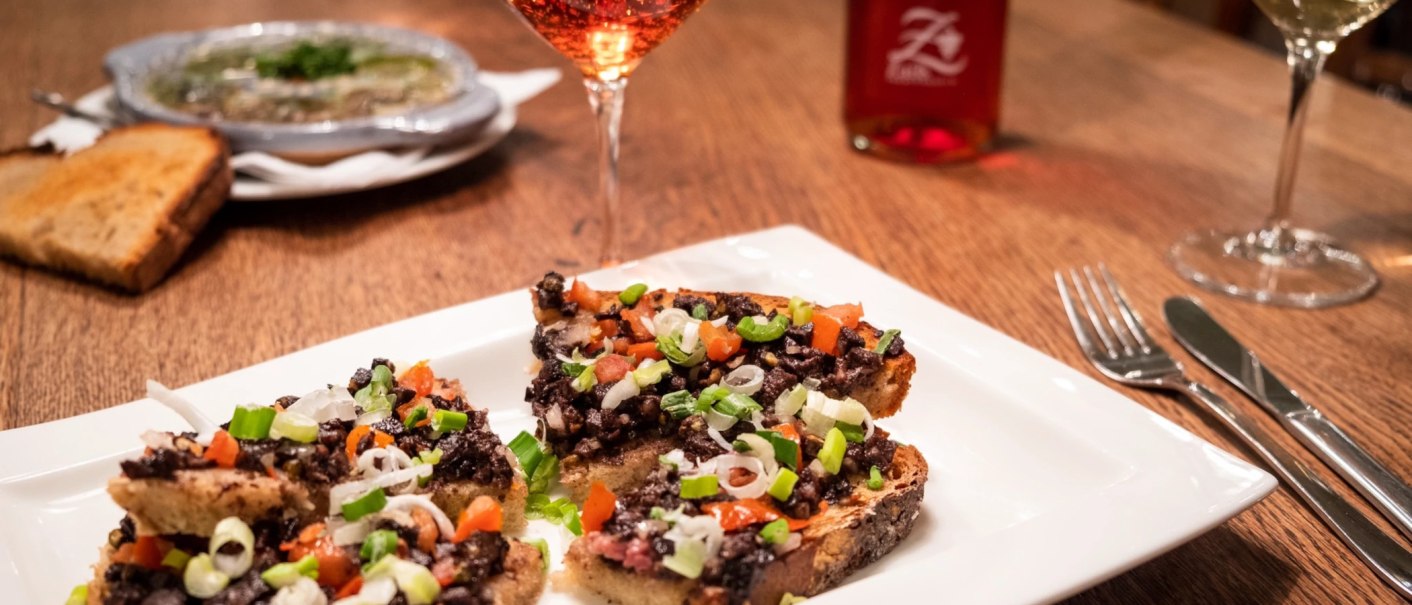 Bruschetta mit Olivenpaste und Gem&uuml;se auf einem wei&szlig;en Teller, daneben ein Glas Ros&eacute;wein auf einem Holztisch., &copy; Weingut Zai&szlig;