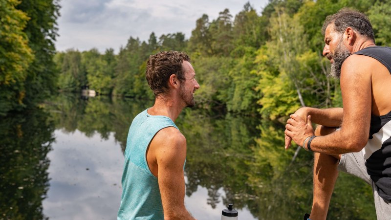 Two men sit on the shore of a lake, surrounded by green trees, and talk. One of them is holding a water bottle., &copy; SMG, Martina Denker