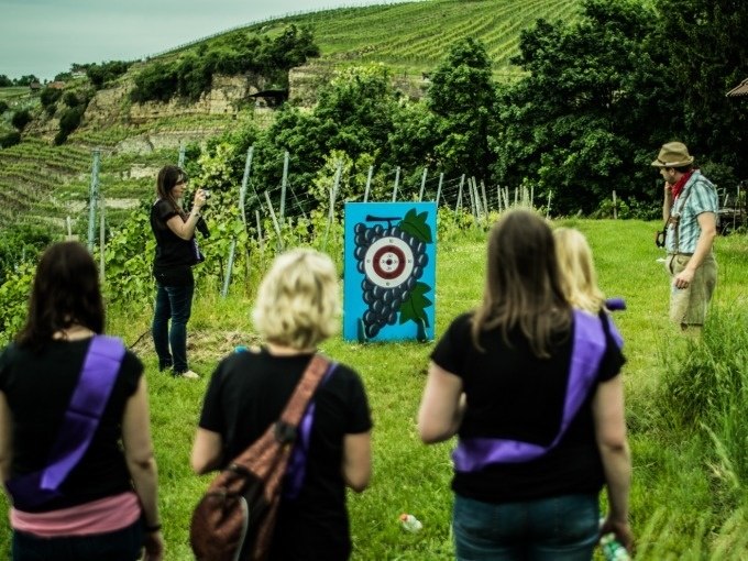 Menschen in einem Weinberg betrachten ein Ziel mit Traubenmotiv. Eine Person fotografiert, andere tragen lila Schärpen., © Cool-Tours StattReisen Menschen in einem Weinberg betrachten ein Ziel mit Traubenmotiv. Eine Person fotografiert, andere tragen lila Schärpen., © Cool-Tours StattReisen