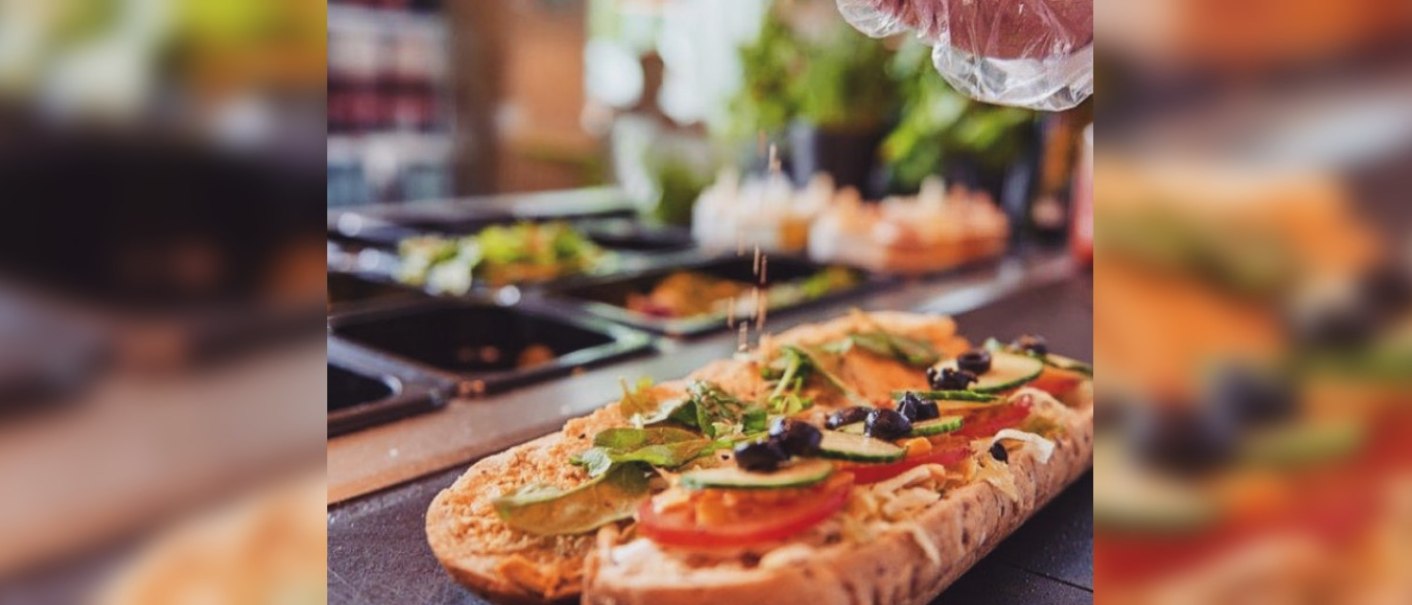 Ein Sandwich wird mit frischem Gemüse und Oliven belegt. Eine Hand streut Gewürze darüber. Im Hintergrund sind Zutaten in Behältern zu sehen., © FreshSub