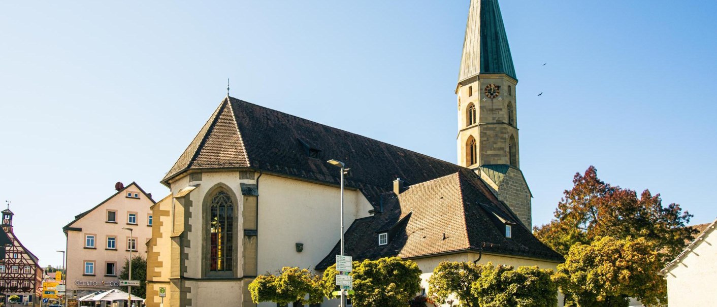 A church with a pointed tower in Gaildorf, surrounded by trees and a half-timbered house in the background under a clear sky., © Stuttgart-Marketing GmbH, Sarah Schmid