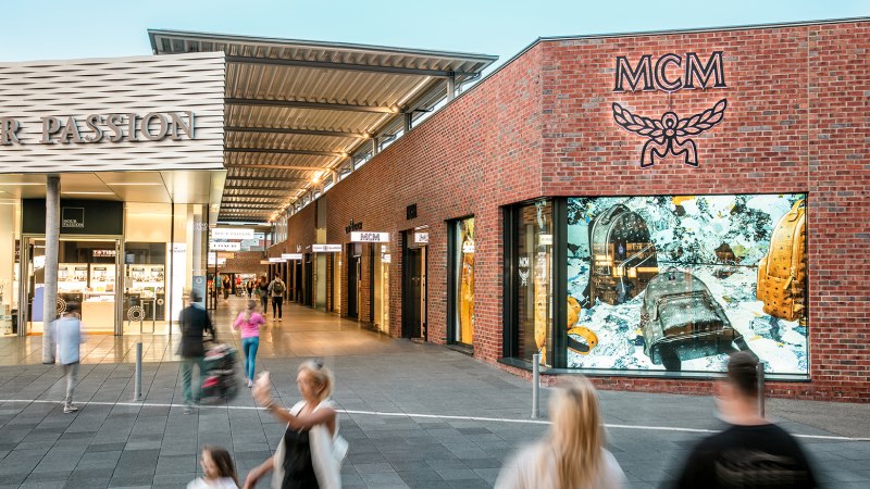 Einkaufsstraße in der Outletcity Metzingen mit Geschäften wie MCM. Menschen schlendern vorbei, einige sind in Bewegung unscharf., © OUTLETCITY AG