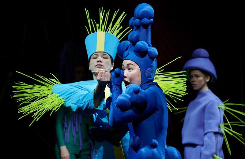 Drei Personen in bunten, fantasievollen Kostümen auf einer Bühne. Eine trägt ein blaues Kostüm mit gelben Details, eine andere ein blaues mit Kugeln, die dritte ein lila Outfit., © Württembergische Staatstheater Stuttgart