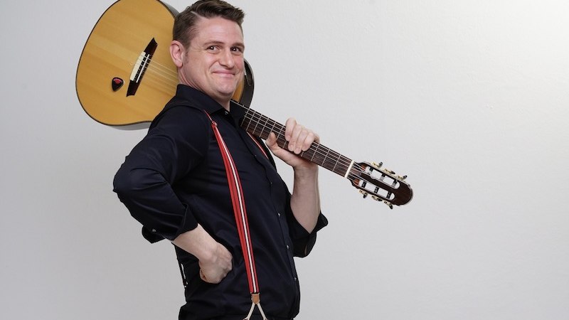 Ein Mann tr&auml;gt eine Gitarre auf der Schulter und l&auml;chelt. Er tr&auml;gt ein schwarzes Hemd und rote Hosentr&auml;ger. Der Hintergrund ist schlicht wei&szlig;., &copy; Theaterhaus Stuttgart e.V.