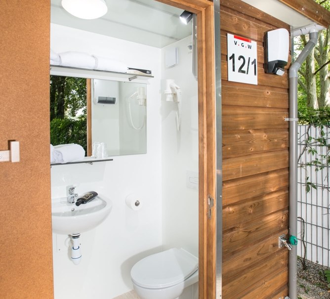 Kleines Badezimmer in einem Holzgebäude mit Toilette, Waschbecken und Spiegel. Außenansicht mit geöffneter Tür, umgeben von Bäumen., © Remsfässer