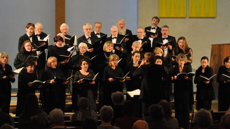 Ein Chor in schwarzer Kleidung singt in einer Kirche, dirigiert von einer Person. Die Sänger halten Notenblätter und stehen vor einem Publikum., © Gertrud Eisenhardt Ein Chor in schwarzer Kleidung singt in einer Kirche, dirigiert von einer Person. Die Sänger halten Notenblätter und stehen vor einem Publikum., © Gertrud Eisenhardt