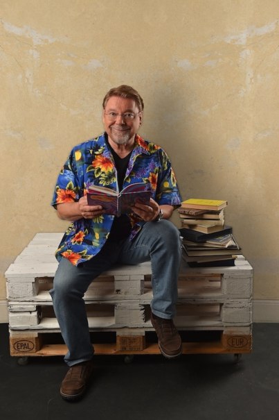 A person in a colorful Hawaiian shirt sits on a white pallet and reads a book. Next to her is a pile of books., &copy; Renitenztheater Stuttgart e.V.