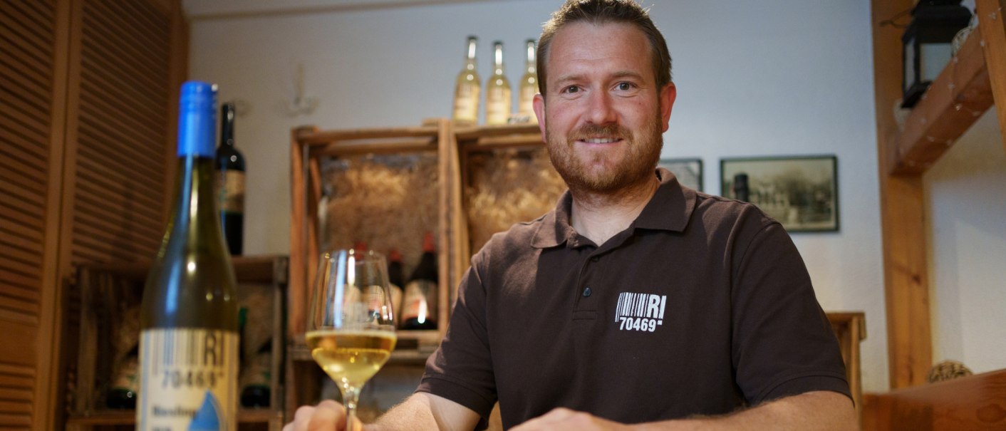 Ein Mann in einem Weinkeller lächelt in die Kamera und hält ein Glas Weißwein. Im Hintergrund sind Weinflaschen zu sehen., © 70469R! Ein Mann in einem Weinkeller lächelt in die Kamera und hält ein Glas Weißwein. Im Hintergrund sind Weinflaschen zu sehen., © 70469R!