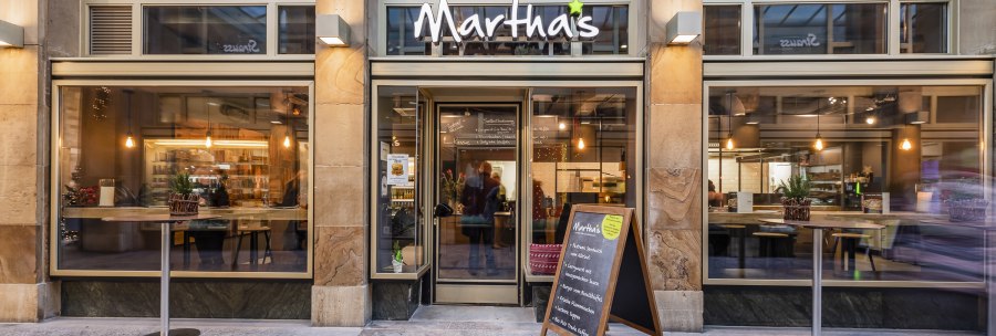 Eingang des Cafés Martha's mit großen Fenstern, Tischen und einer Speisekarte im Freien. Innen sind Lichter und Dekorationen sichtbar., © Matha's, Stuttgart