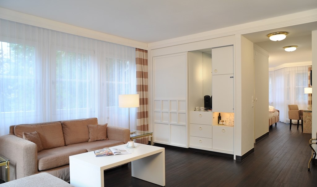 Bright living area of a junior suite with beige sofa, white coffee table and view of the sleeping area. Large windows with curtains., &copy; relexa Waldhotel Schatten
