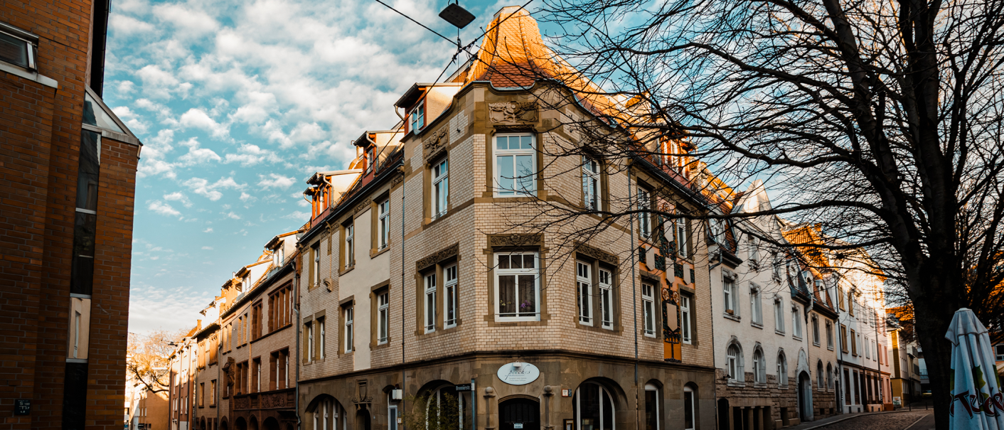 Eckgebäude mit roten Ziegeldächern, verziertem Mauerwerk und großen Fenstern. Ein Baum ohne Blätter steht daneben, blauer Himmel im Hintergrund., © Stuttgart-Marketing GmbH, Sarah Schmid