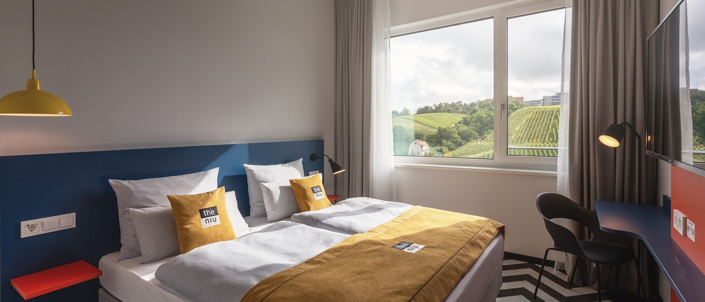 A modern hotel room with a double bed, yellow accents, and a large window overlooking vineyards., © the niu Form - Novum Hospitality A modern hotel room with a double bed, yellow accents, and a large window overlooking vineyards., © the niu Form - Novum Hospitality