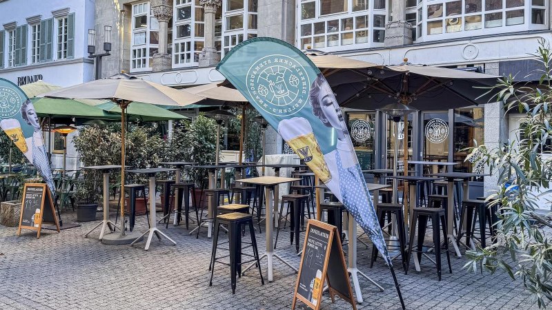 The outdoor area of the Dinkelacker Stehausschank, featuring bar tables, umbrellas, and beer banners in front of a historic building., &copy; SMG Stuttgart Marketing GmbH - Ellen Haeffner